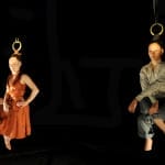 Two aerial performers with their hair tied to metal rings hang suspended in the air against a dark backdrop. One wears a flowing orange dress, while the other is dressed in a grey suit, both appearing poised and weightless.