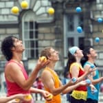 eight jugglers wearing brightly coloured costumes all in a straight line, juggle yellow, blue and pink balls in the air against the outside brickwork of Somerset House courtyard Free Time | Somerset House
