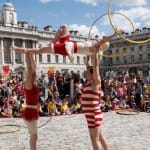 three acrobats dressed in red and white old-fashioned swimwear perform to a large outdoor audience in the Somerset House courtyard, two acrobats hold up the third who twirls two gold hoops, one in each hand Free Time | Somerset House