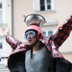 A performer in a dazzling, sequined jacket stands in a joyful pose with both hands up, wearing a bowl on their head against a backdrop of buildings.