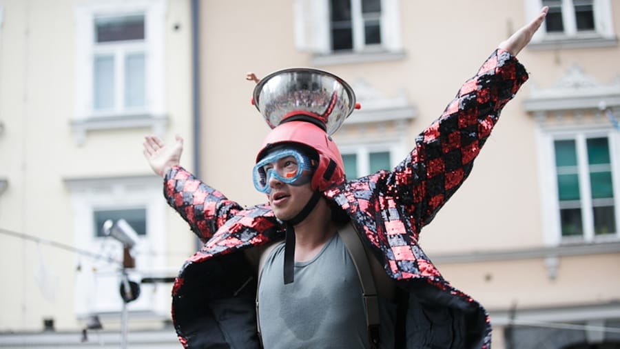 A performer in a dazzling, sequined jacket stands in a joyful pose with both hands up, wearing a bowl on their head against a backdrop of buildings.