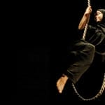 A performer in head to toe black costume swings gracefully on a rope against a dark backdrop, creating a dramatic and mysterious atmosphere What If…? | Layla Rosa ©