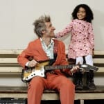 A musician in an orange suit sits on a bench, playing a guitar, while a child in a pink dress stands beside them, both smiling and looking at each other.