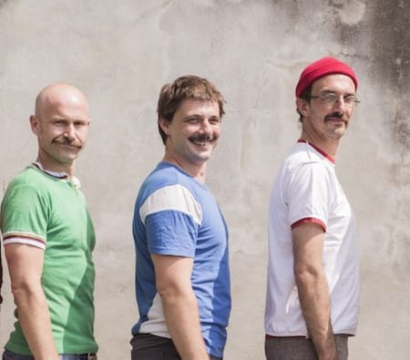 Five men stand sideways, in descending height order, each wearing a different coloured shirt, against a textured, light-coloured wall.