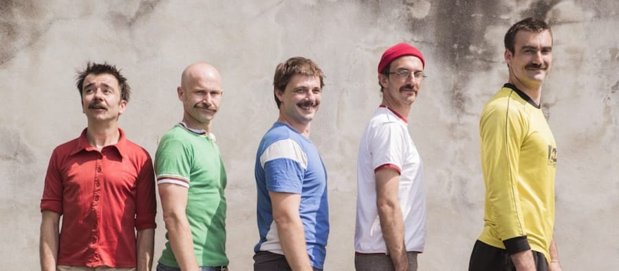 Five men stand sideways, in descending height order, each wearing a different coloured shirt, against a textured, light-coloured wall.
