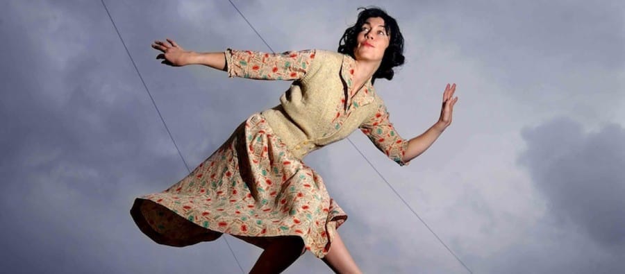 A woman in a vintage floral dress striking a dynamic pose on a rooftop, appearing as if caught mid-motion, with a moody sky behind her.