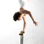 A acrobat performs an impressive handstand on a wooden apparatus against a minimalistic, white wall background.