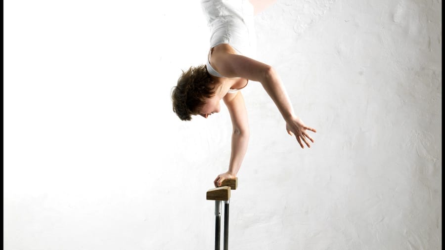 A acrobat performs an impressive handstand on a wooden apparatus against a minimalistic, white wall background.