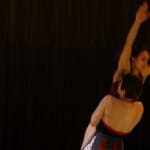 Two performers are being lifted into the air in a dramatic pose, set against a dark backdrop.