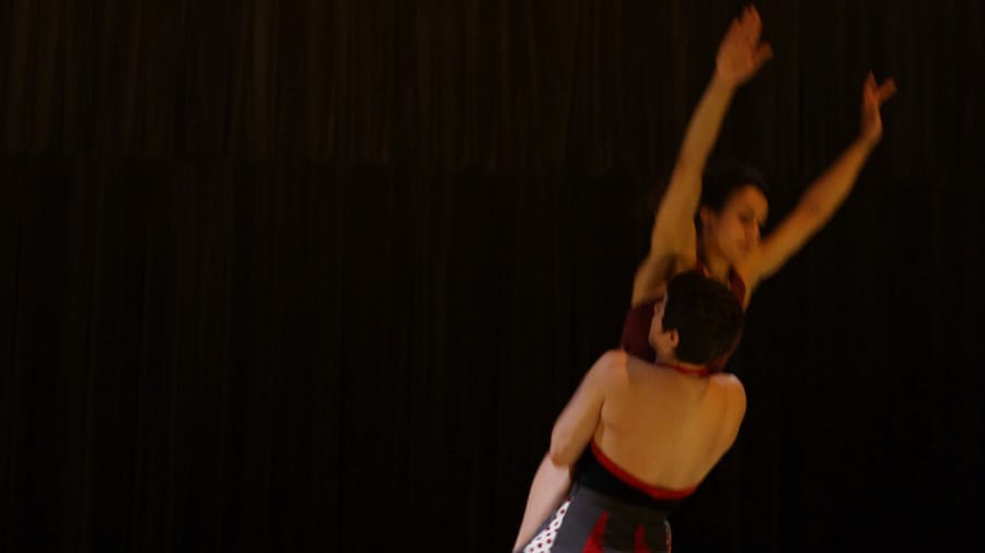 Two performers are being lifted into the air in a dramatic pose, set against a dark backdrop.