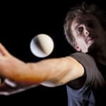 A person with brown hair, dressed in a black shirt, gracefully juggles two white balls against a deep black background.