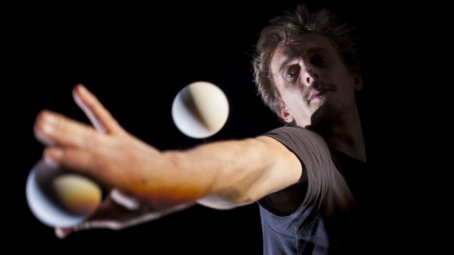 A person with brown hair, dressed in a black shirt, gracefully juggles two white balls against a deep black background.