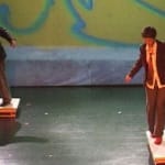 Two performers in formal attire balance on wooden platforms against a colourful backdrop, showcasing a dynamic dance movement.