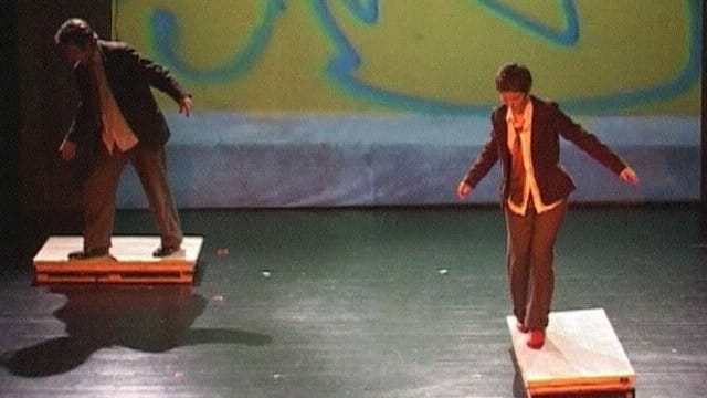 Two performers in formal attire balance on wooden platforms against a colourful backdrop, showcasing a dynamic dance movement.