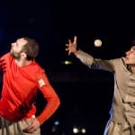Two performers in vibrant attire - one dressed in red and the other in light brown - engage in a dynamic juggling act, illuminated by bright stage lights.