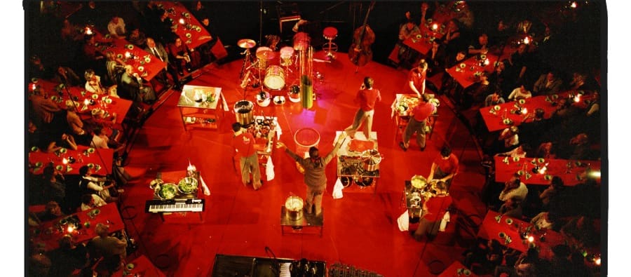 Aerial view of a vibrant performance on a red circular stage surrounded by audience tables. Performers in red outfits engage in an energetic cooking show, using various kitchen instruments while interacting with the crowd, creating a lively dining atmosphere. Belgian company Laika give you Patatboem