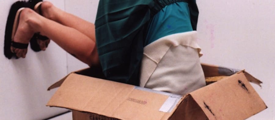 A person in a teal skirt and white sweater is upside down in a cardboard box, with their head hidden inside and legs sticking out, creating a surreal visual effect.