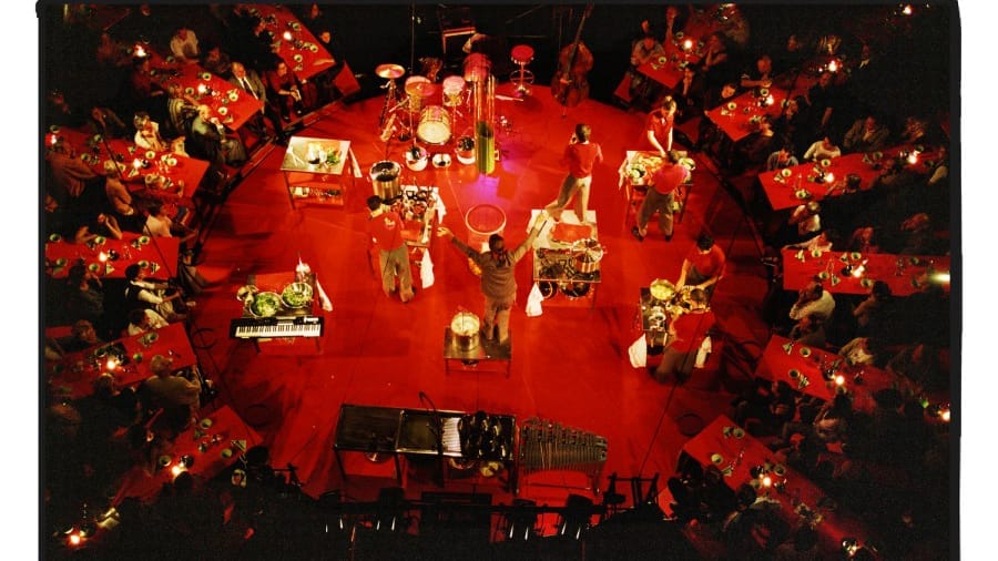 Aerial view of a vibrant performance on a red circular stage surrounded by audience tables. Performers in red outfits engage in an energetic cooking show, using various kitchen instruments while interacting with the crowd, creating a lively dining atmosphere.