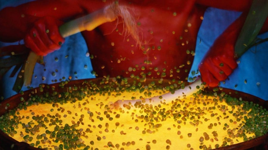 A performer energetically stirs a large, illuminated pot, causing green peas to fly into the air. The dramatic lighting highlights the movement and intensity of the action.