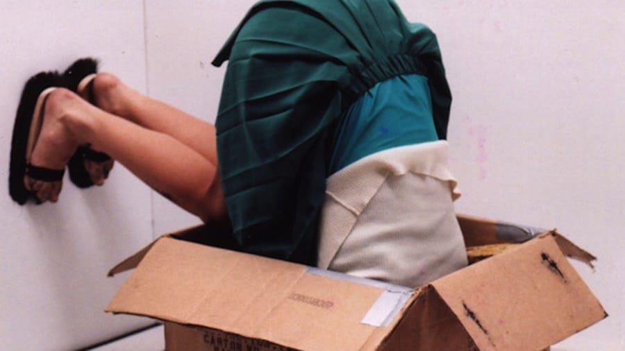 A person in a teal skirt and white sweater is upside down in a cardboard box, with their head hidden inside and legs sticking out with their feet against the wall, creating a surreal visual effect Casio Tone © Mónica Lapa, Joseph Fabião