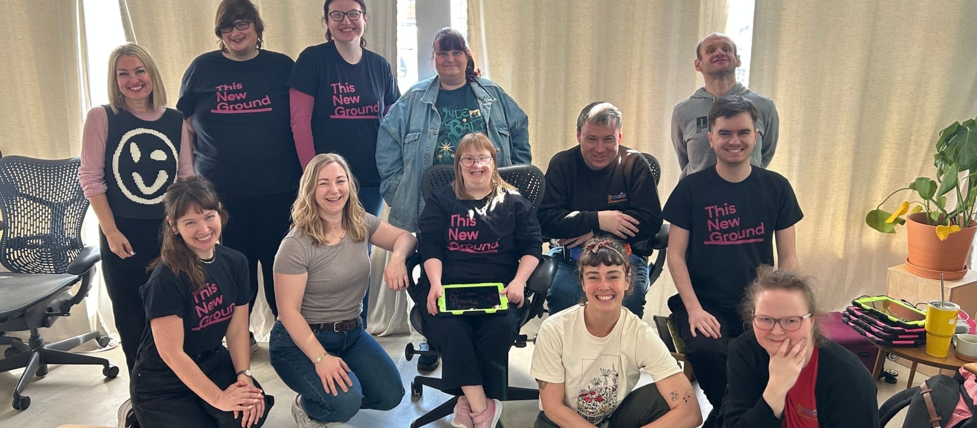 ten members of The New Ground organisation with Lucy Babb from Crying Out Loud and Tilly Lee-Kronick all posing for the camera in a curtained room, some of the organisation are wearing The New Ground black t-shirts with pink writing Groundwork project | Portsmouth