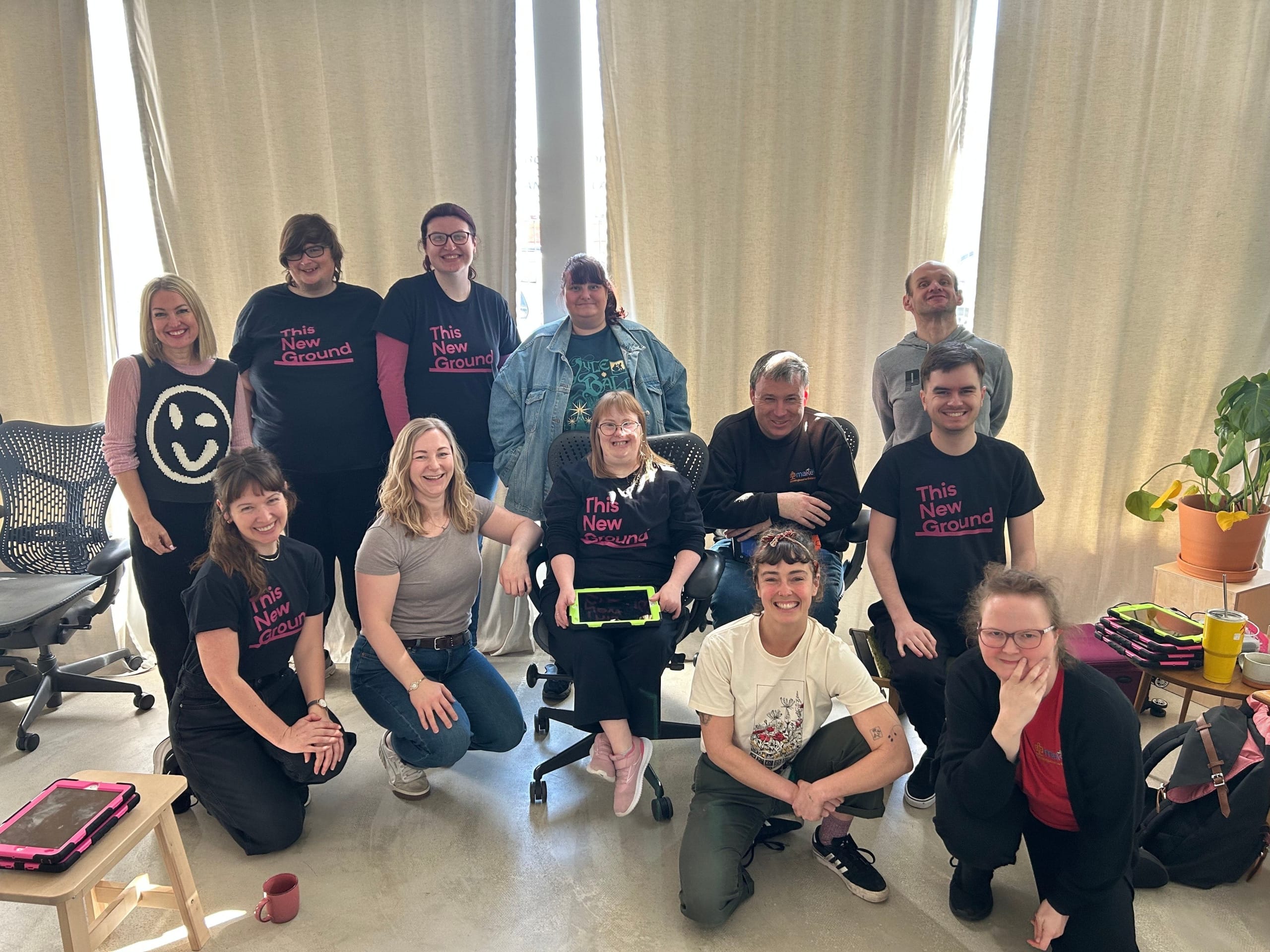 ten members of The New Ground organisation with Lucy Babb from Crying Out Loud and Tilly Lee-Kronick all posing for the camera in a curtained room, some of the organisation are wearing The New Ground black t-shirts with pink writing Groundwork project | Portsmouth