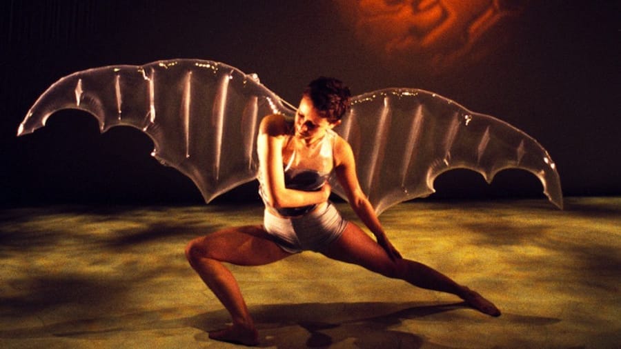 A dancer in a dynamic pose wears striking clear bat-like wings, set against a warm, atmospheric backdrop.