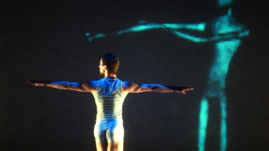 A dancer in a white outfit stands on stage with arms extended and back turned, illuminated against a dark background with a blue holographic silhouette.