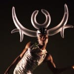 A dancer in a white sleeveless camisole poses dramatically, wearing an oversized headpiece resembling abstract horns, against a dark background.