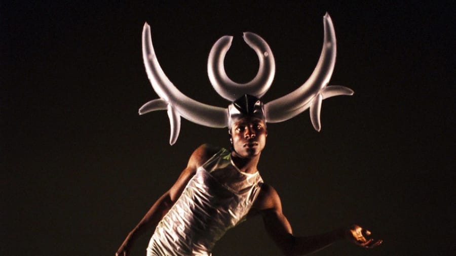 A dancer in a white sleeveless camisole poses dramatically, wearing an oversized headpiece resembling abstract horns, against a dark background.