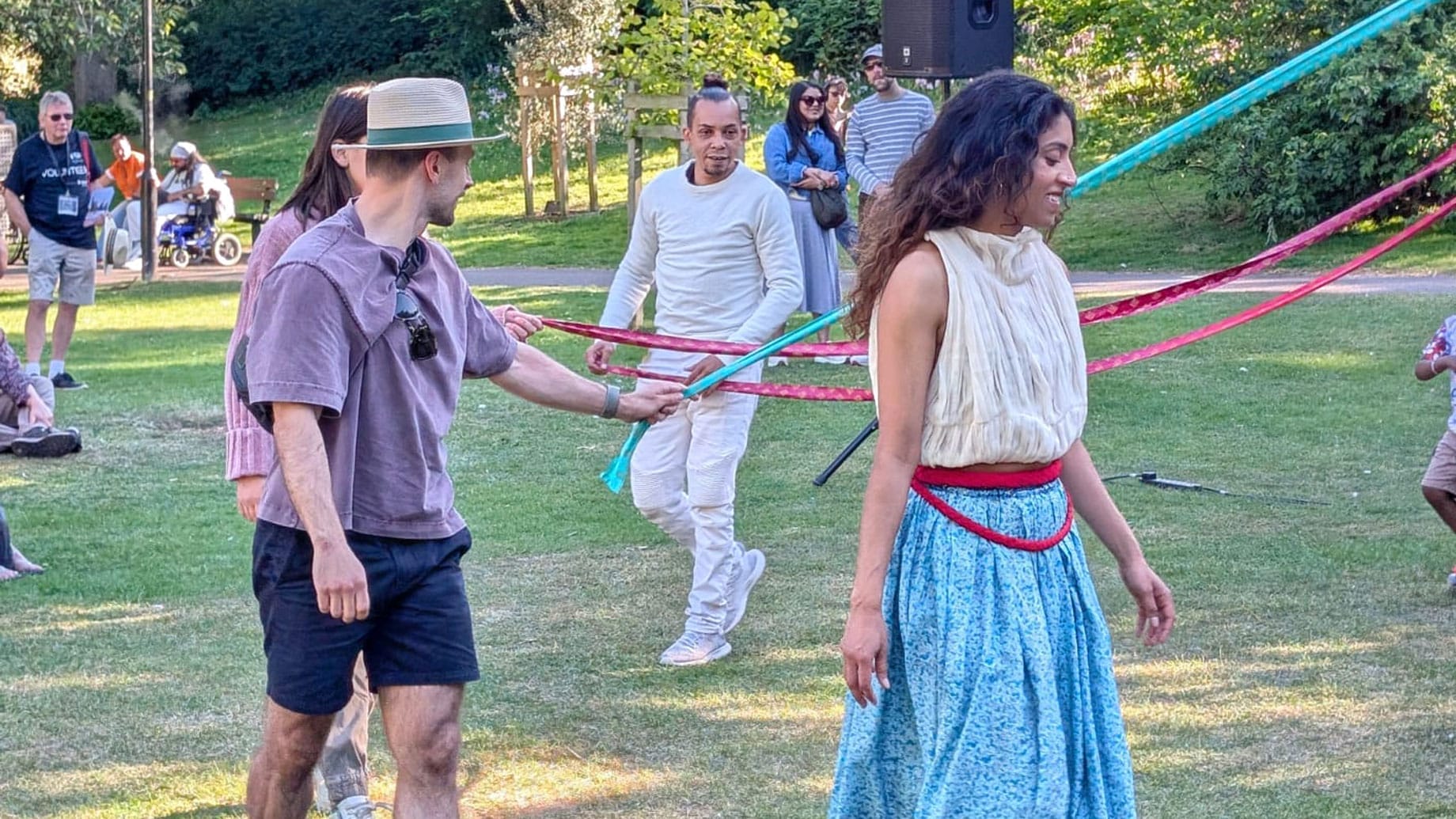 One performer dressed in a blue and white costume, and three others move in harmony around a May pole, in an outdoor green park Solent Artist Inspiration Trip