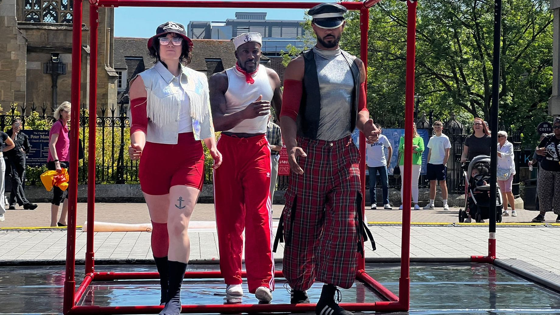 Three performers all dressed in red and white casual costumes, wearing hats and sunglasses move in harmony on an outdoor stage against a cathedral and blue sky with an audience watching Solent Artist Inspiration Trip