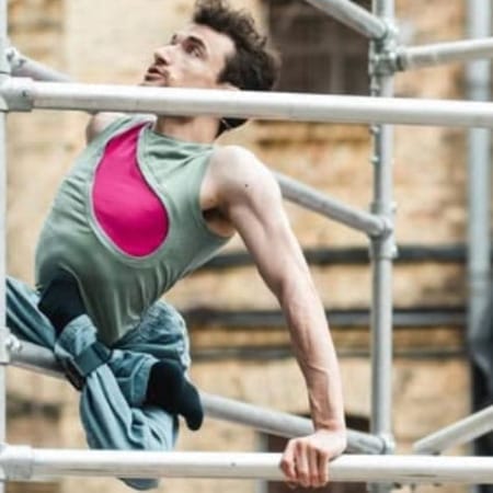 an acrobat with short brown hair wearing a pink and green shirt with blue trousers climbs an outdoor scaffold, their legs are crossed and tied beneath them, an audience watches in front of a brick building Circuliacija Carousel artists Head Over Wheels