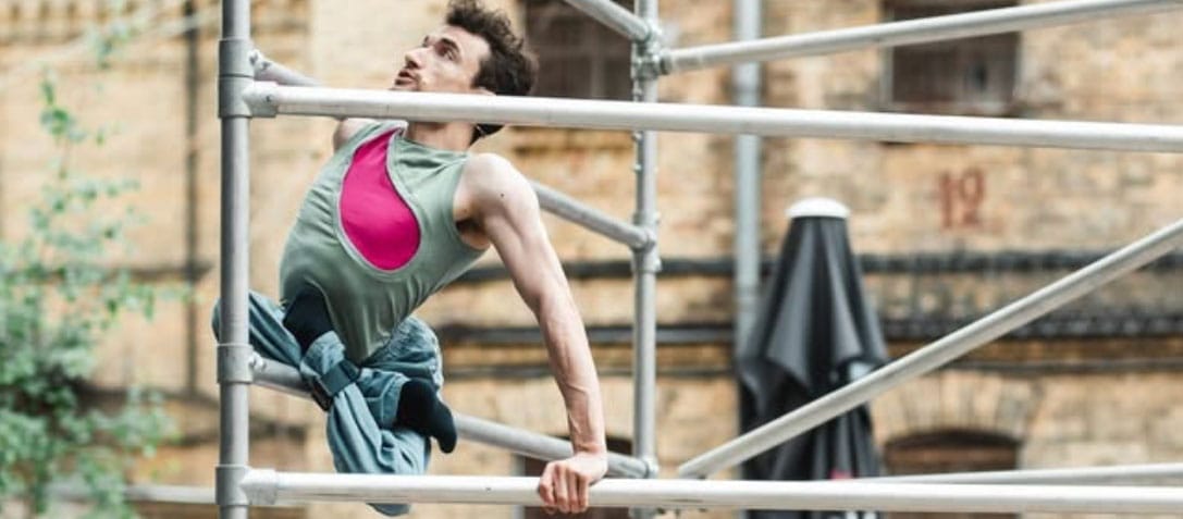 an acrobat with short brown hair wearing a pink and green shirt with blue trousers climbs an outdoor scaffold, their legs are crossed and tied beneath them, an audience watches in front of a brick building Circuliacija Carousel artists Head Over Wheels