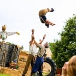 three performers and one scarecrow, in an outdoor setting, two throw up into the air the third performer, they all wear autumnal colours with either braces or dungarees Farm Yard Circus Masterclass