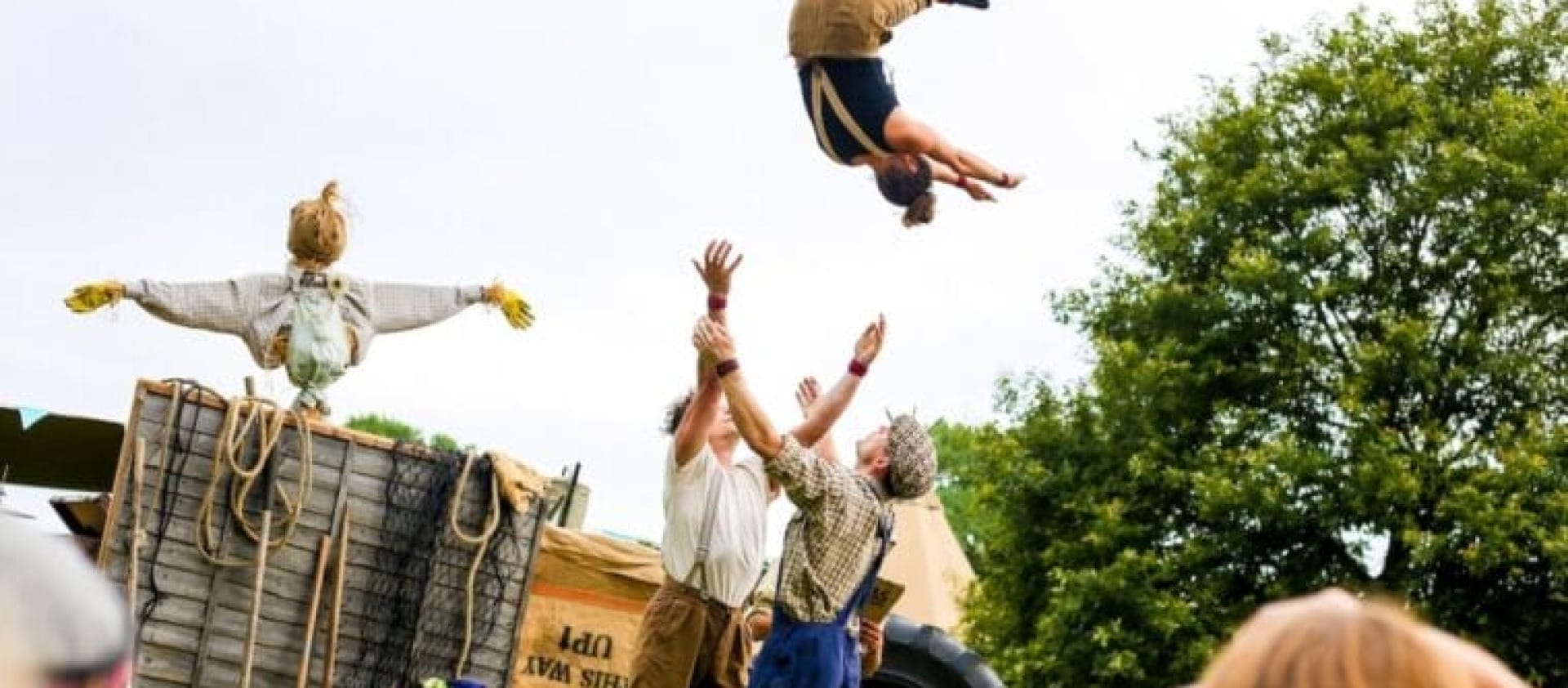 three performers and one scarecrow, in an outdoor setting, two throw up into the air the third performer, they all wear autumnal colours with either braces or dungarees Farm Yard Circus Masterclass
