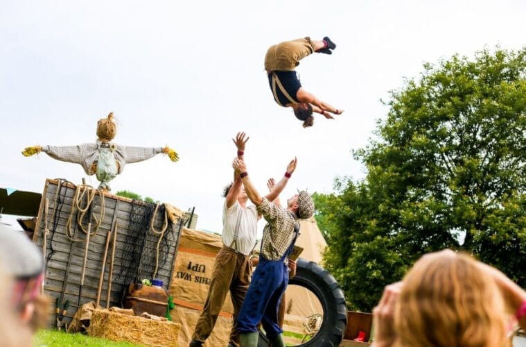 three performers and one scarecrow, in an outdoor setting, two throw up into the air the third performer, they all wear autumnal colours with either braces or dungarees Farm Yard Circus Masterclass