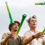 two performers in an outdoor scene juggle four prop leaks, they both wear autumnal colours, one wears a brown hat the other has unruly curly hair Farm Yard Circus | Leeks