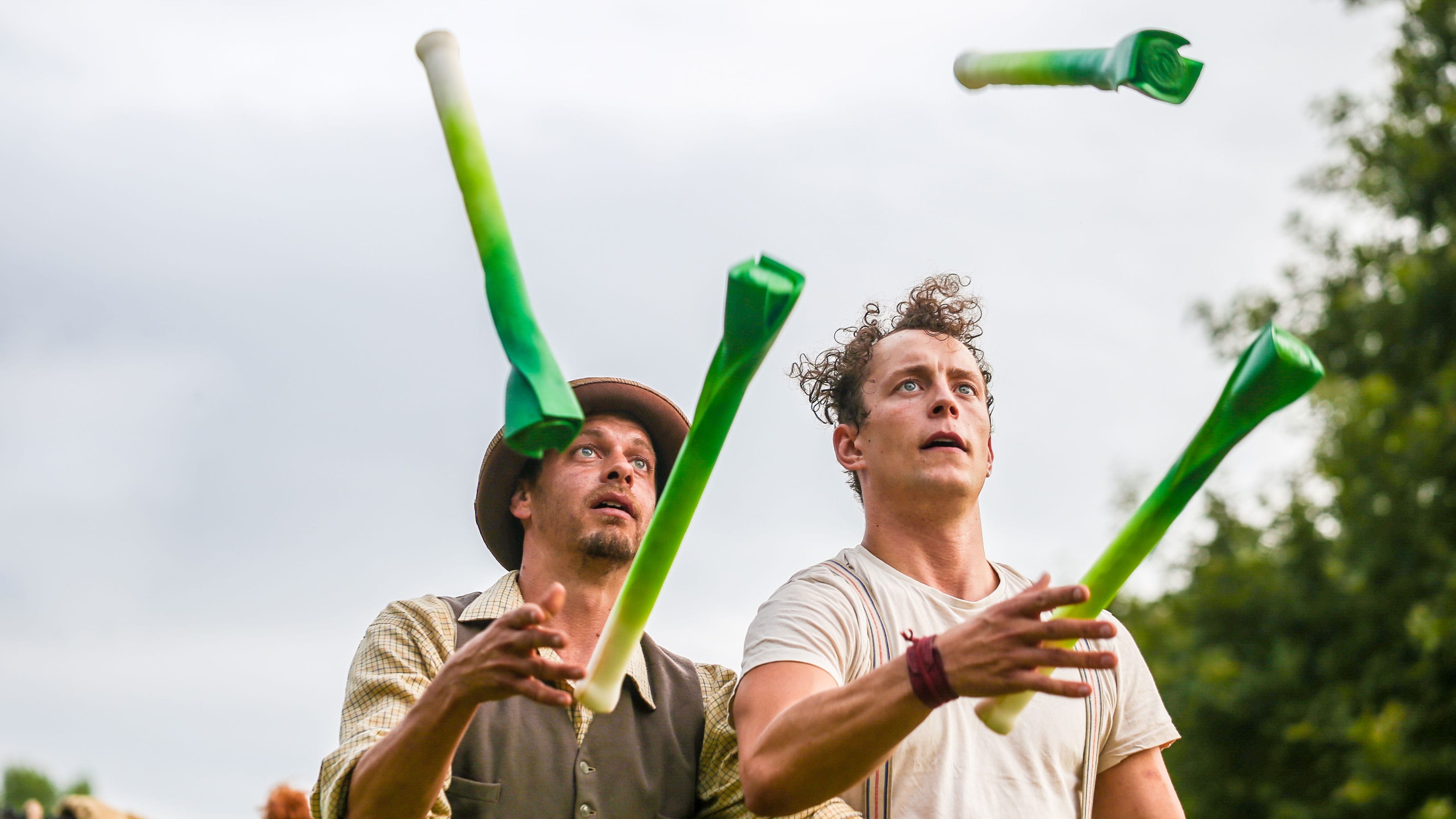 two performers in an outdoor scene juggle four prop leaks, they both wear autumnal colours, one wears a brown hat the other has unruly curly hair Farm Yard Circus | Leeks