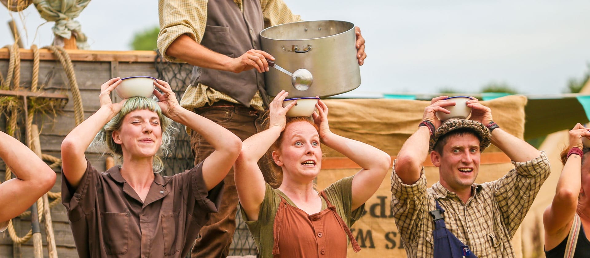 six performers and one scarecrow, in an outdoor setting, one ladles from a large cooking pot soup into small bowls held by the other five on their heads, they all wear autumnal colours with either braces or dungarees Farm Yard Circus | Soup