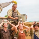 six performers and one scarecrow, in an outdoor setting, one ladles from a large cooking pot soup into small bowls held by the other five on their heads, they all wear autumnal colours with either braces or dungarees Farm Yard Circus | Morning Farm Yard Circus | Soup