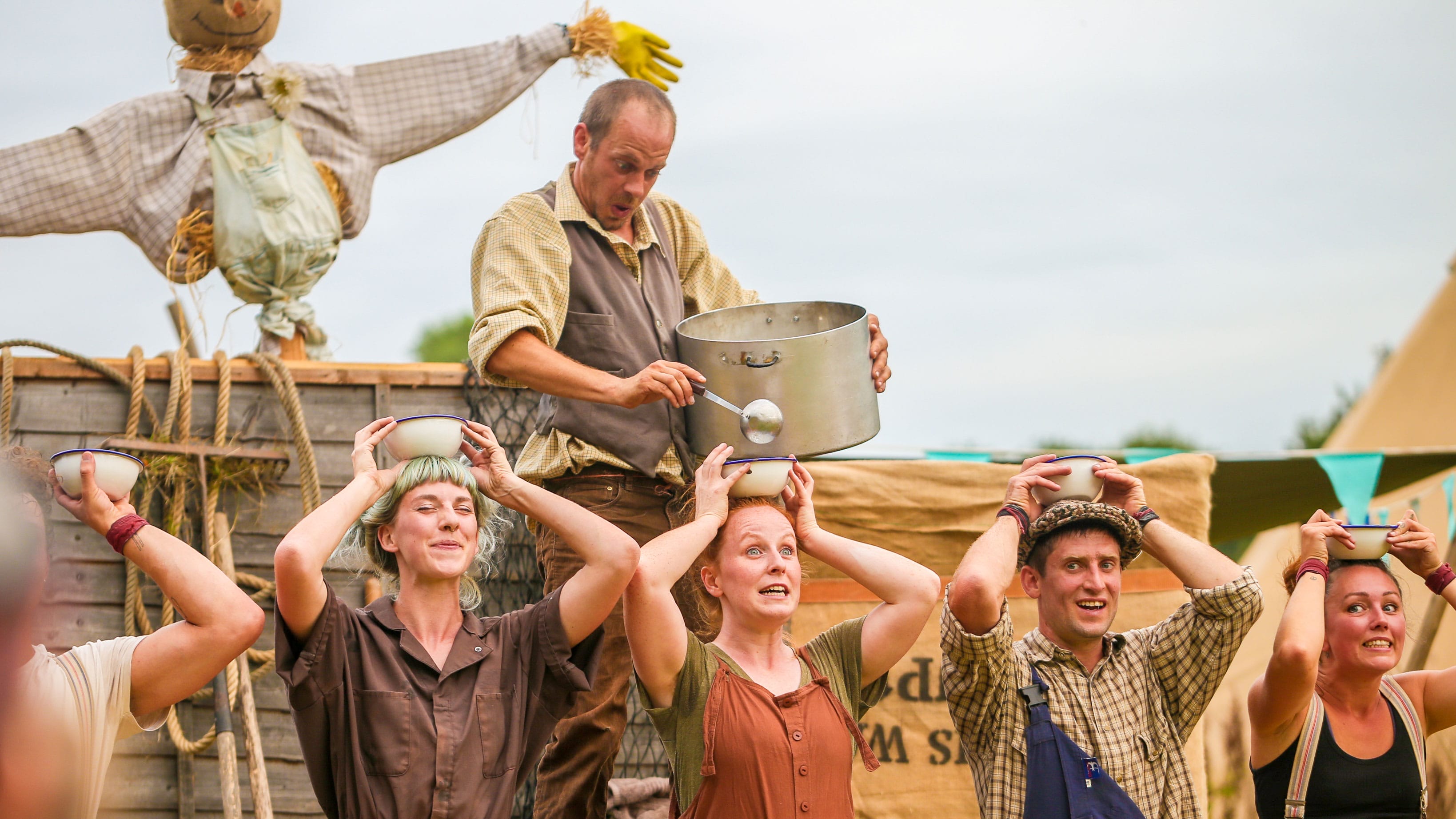 six performers and one scarecrow, in an outdoor setting, one ladles from a large cooking pot soup into small bowls held by the other five on their heads, they all wear autumnal colours with either braces or dungarees Farm Yard Circus | Morning Farm Yard Circus | Soup