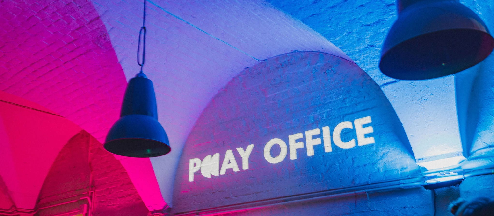 the words PLAY OFFICE are projected onto a white brick vaulted wall space, the accompanying lighting is pink and blue with two hanging light fittings in the foreground Play Office | Portsmouth Historic Dockyard
