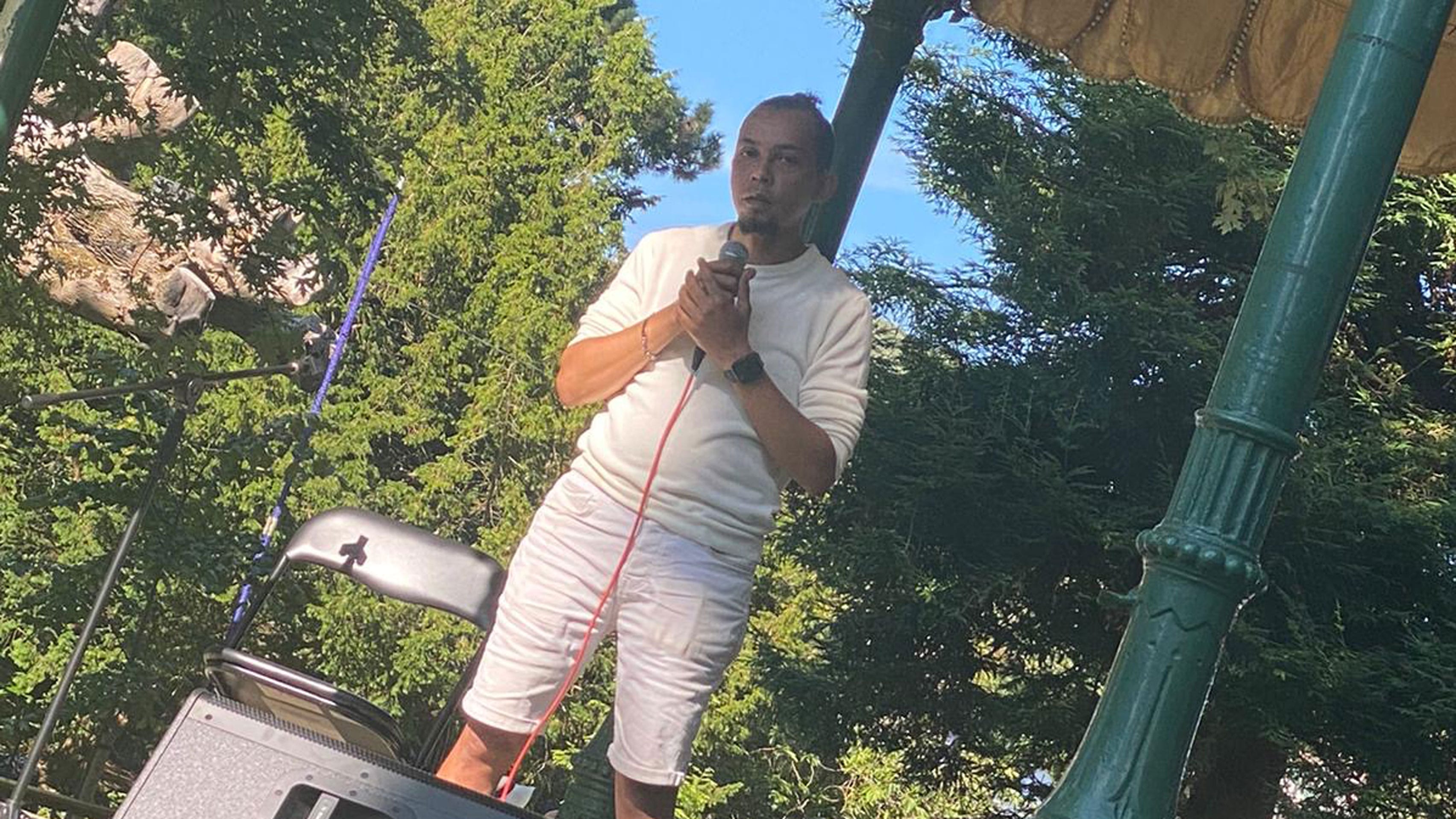 a poet stands on an outdoor festival on bandstand stage holding a microphone, with a small brown beard and wearing a white t-shirt and white shorts Artist Inspiration Trip | Ventnor 2024