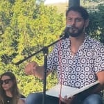 a poet sits on an outdoor festival stage in front of a microphone, with a brown beard and a flowery patterned shirt and blue jeans Artist Inspiration Trip | Ventnor 2024