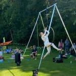 an aerial hoop performer in a white body suit sits on a hoop hanging from a tripod at an outdoor festival with an audience watching while sitting on the grass Artist Inspiration Trip | Ventnor 2024