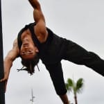 a performers holds their body horizontally on an outdoor Chinese pole, with a small beard and moustache they are wearing a pair of black trousers and vest, they face the camera, there are palm trees in the background Human Flag | Brahim Ech-Chibi