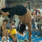 a performers holds their body horizontally on a Chinese pole, wearing a pair of black trousers and no other clothing, they face away from the camera Human Flag | Brahim Ech-Chibi