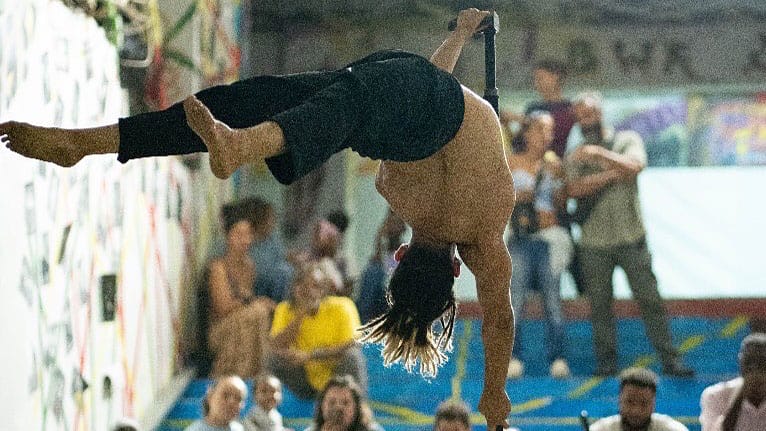 a performers holds their body horizontally on a Chinese pole, wearing a pair of black trousers and no other clothing, they face away from the camera Human Flag | Brahim Ech-Chibi