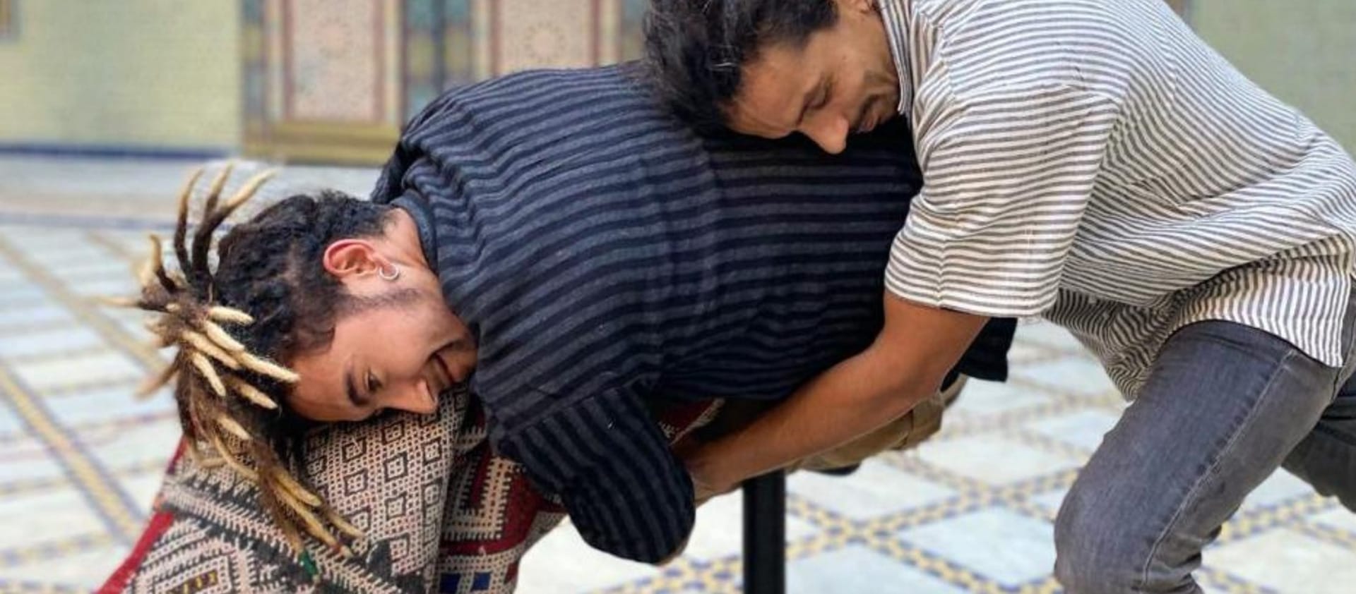 two performers train in a indoor rehearsal studio laid with patterned carpets, one performer is embracing the other resting their head on their back, the other performer holds a rolled up carpet mimicking the embrace, both have facial hair and wear striped shirts and dark jeans Zarbia | Company Zid and Brahim Ech-Chibi Zarbia work-in-progress